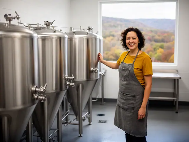 A smiling brewer in a gray apron beside new stainless-steel conical fermenters, wooded hills outside.